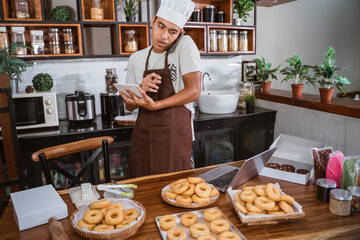 Asian Chef calls using a smartphone while matching notes on a donut order from a notebook with a laptop, working on making homemade donuts