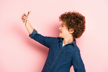 side view of curly kid in denim shirt taking selfie on smartphone on pink