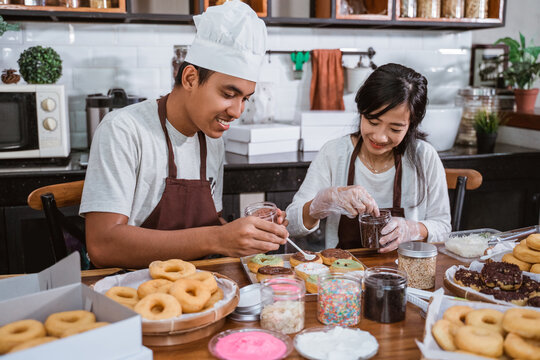 A married couple who sit wearing aprons while making donuts and decorating donuts with chocolate in the kitchen