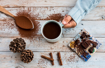 A female hand holding a blue cup filled with dark, hot, creamy and thick chocolate on a wooden table as a background with dark chocolate pieces and spoon with cocoa powder