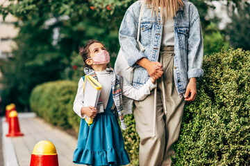 The kid wearing a protective face mask going to the school with her mother during coronavirus. Happy little girl with a backpack and folder goes to the school with her mom.