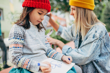 Side view of the cute little girl in the red cap is sitting outdoor and drawing on paper. Daughter enjoying the time together with her mother outside. Mom and daughter share love. Mother's day.