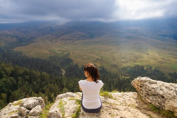 Naklejka premium Rear view of a young woman sitting at the top of a mountain, looking into the distance on the top of mountain. Background is mountains and cloudy sky.