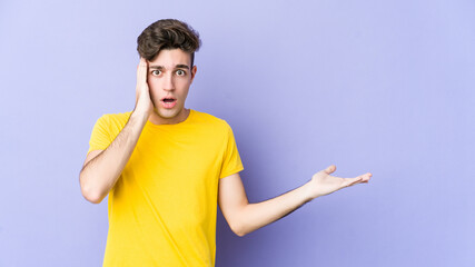 Fototapeta premium Young caucasian man isolated on purple background impressed holding copy space on palm.