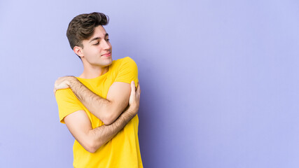 Young caucasian man isolated on purple background hugs, smiling carefree and happy.