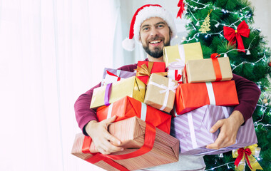 Obraz na płótnie Canvas Handsome excited bearded young man in santa hat with many presents in hands is posing on christmas tree background