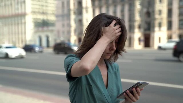portrait of a sad middle-aged brunette in a green dress. she is walking along a city street against the background of passing cars, holding her forehead, checking phone
