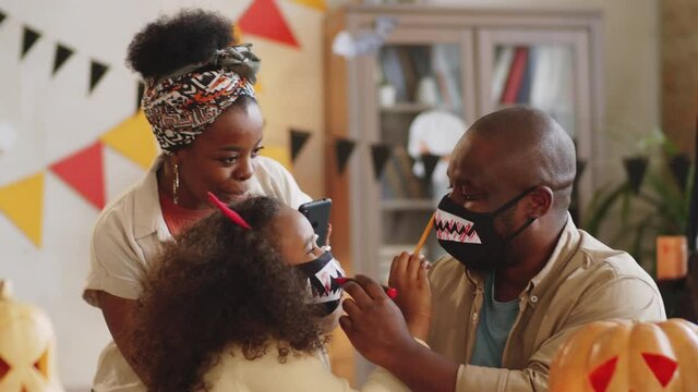 Little African American girl and her father drawing blood on vampire teeth masks with felt tip pens while cheerful mother taking picture with smartphone during Halloween preparation at home