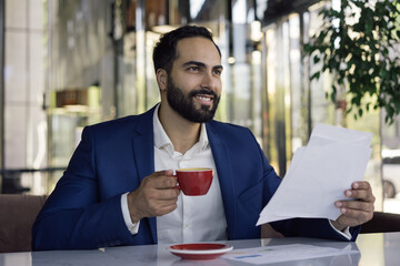 Portrait of young handsome arabic manager working with documents, sitting at workplace. Smiling middle eastern businesswoman reading contract, planning start up, holding cup of coffee in modern office