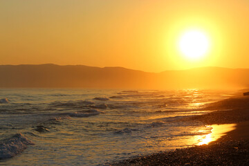 Big yellow sun setting over the sea. Waves hit the pebble beach. Mountains in the background