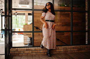 Fashion portrait of african american woman in long dress, boots and sunglasses indoor.