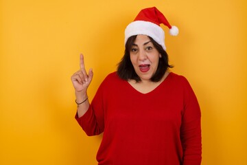 Close up portrait of pleasant looking middle aged woman wearing Santa hat has clever expression, raises one finger, remembers herself not to forget tell important thing. Indoors