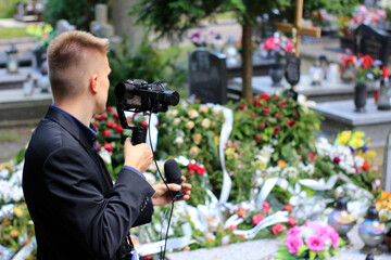 Elegant man in suit is recording grave with cross full of flowers. Funeral live streaming on cementary. Cameraman handing a microphone and camera on a gimbal.