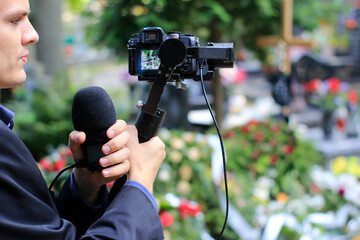 Elegant man in suit is recording grave with cross full of flowers. Funeral live streaming on cementary. Cameraman handing a microphone and camera on a gimbal.