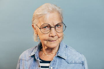 Upset unsatisfied elderly woman with white short hair in round glasses wearing denim jacket and striped shirt. Irritated old lady looking at camera seriously. Isolated over blue background.
