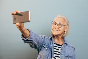 Happy smart old lady in blue jeans jacket, black and white striped shirt smiles and takes selfie holding mobile phone at arm length. Online communication for elderly people concept.