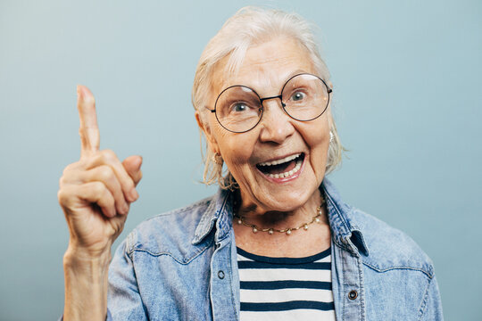 Excited positive old woman opens laughs opening her mouth and points up with her index finder. Happy elderly lady wearing smart round glasses and casual clothes isolated over light blue background.
