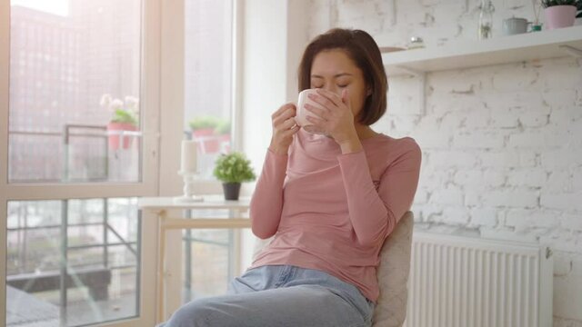 Young Asian Woman Relaxing And Holding Cup Enjoying Aroma Looking Through Window Enjoying Peaceful Moment At Home, Girl Sitting In Comfortable Chair In Cozy Apartment Dreaming, Pleasant Leisure Day