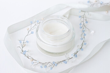 Glass jar of a cosmetic cream on a white background. White ribbon and blue bijouterie on a table