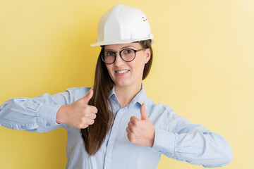 Portrait of smiling beautiful tatar woman engineer in construction helmet and glasses showing thumbs up isolated on yellow background