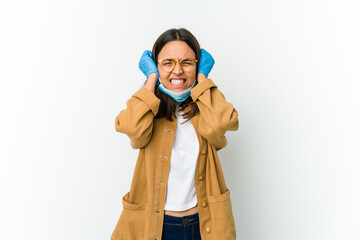 Young latin woman wearing a mask to protect from covid isolated on white background covering ears with hands trying not to hear too loud sound.