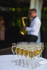 Table with glasses of champagne, a saxophonist plays in the background. Against the background of a blurry saxophonist with saxophone, the atmosphere of the holiday cocktail reception with champagne.