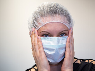 Young girl in a medical surgical blue color mask. On the head is a protective cap or hat. The model is isolated on a light-colored wall background. Quarantine and freedom restriction concept. Close-up