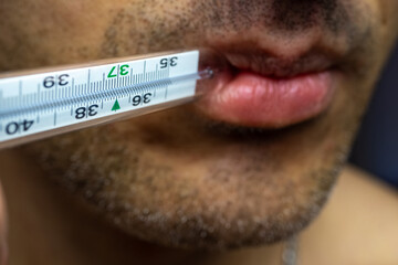 A tanned man holds a glass thermometer in his mouth to measure body temperature. Thermometer between the lips, bristles on the face. The concept of a healthy lifestyle and health care. Close-up.
