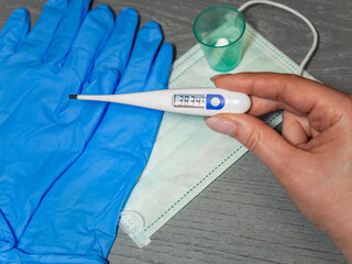 Green medicine mask, pill, white electronic thermometer in woman hand and pair of blue gloves on gray wooden background. The concept of medical supplies or accessories for personal protection.