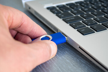 A tanned male hand inserts or connect a blue USB flash drive to laptop computer's USB port. Notebook is on a light gray desk background.  Data storage and modern electronics concept. Close-up.
