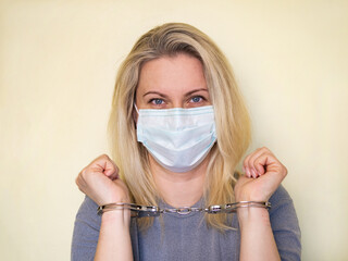 Young girl in a mask with handcuffed hands . Handcuffs are put on raised hands. The model is isolated on a light-colored wall background. Quarantine and freedom restriction concept. Close-up
