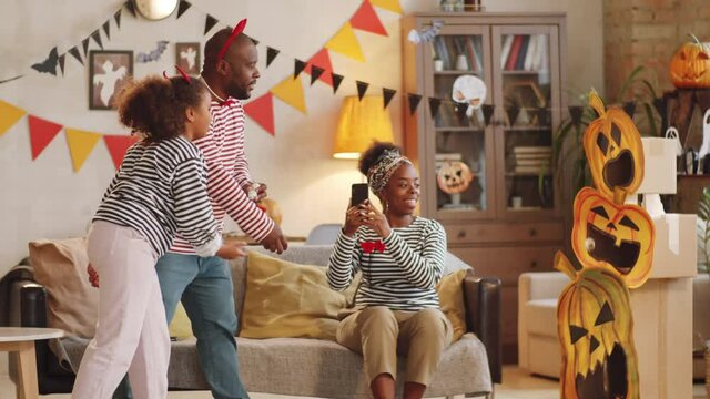 Little black girl and her father throwing balls into boxes with paper jack-o-lanterns while playing at home on Halloween, cheerful mother taking pictures of their game with smartphone