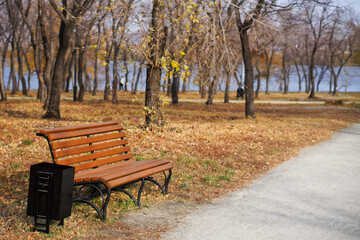 An empty bench next to a path in an autumn park near a lake or river. Change of seasons.