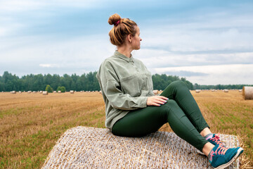 Young european woman sitting on the round bale of hay on the agriculture field in the village