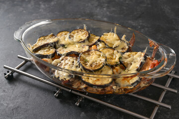 Vegetable gratin with eggplants, tomatoes and bell pepper baked with cheese in a glass casserole dish on a dark stone background