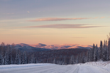 Fototapeta na wymiar The road in the winter mountains in the background. Winter valley panorama with forest and mountain peaks in the background at sunrise. First rays of the sun in the morning