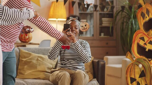 Happy little afro-american girl playing Halloween game and tossing the ball into jack-o-lantern box, jumping with excitement and hugging dad while mother taking picture of them with smartphone