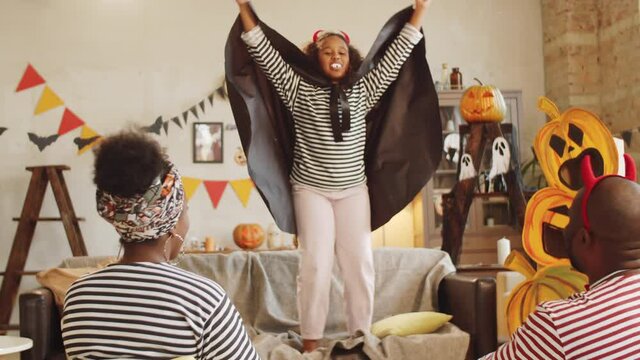 Cute black girl in vampire costume standing on sofa in living room and giving Halloween performance to mother and father, happy parents sitting on floor and clapping hands