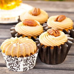 Salted Caramel Bourbon Cupcakes in Black Cupcake Wrappers Topped with Pecan Halves on Wooden Table with Bourbon in Glass