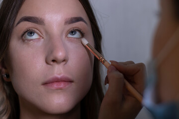 the process of applying makeup to a beautiful brunette by a master in a medical mask. beauty, modeling and cosmetics concept.