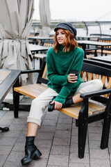 Portrait of a beautiful red-haired girl.  Beautiful red-haired girl in a green sweater and cap sits with a cup of coffee in a cafe on the street.