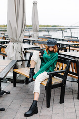 Portrait of a beautiful red-haired girl.  Beautiful red-haired girl in a green sweater and cap sits with a cup of coffee in a cafe on the street.