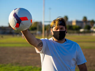 Hombre gordo con tatuajes sujetando un balón mientras viste mascarilla para respetar las medidas...