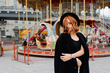 Obraz na płótnie Canvas A beautiful red-haired girl in a black coat and hat stays near the merry-go-round.