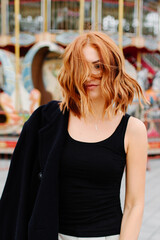 A beautiful red-haired girl in a black coat and hat stays near the merry-go-round.