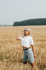 Naklejka na ściany i meble Woman in a white T-shirt and shorts in the field. blonde woman in sunglasses. A beautiful fit tanned girl. Sexy slim model outdoors. . High quality photo