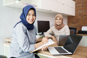 attractive cheerful young muslim business woman working on laptop and smiling while sitting at her desk modern office with her friend at studio