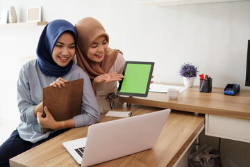 attractive cheerful young muslim business woman working on laptop tablet and smiling while sitting at her desk modern office with her friend at studio