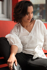 cute young Latin brunette woman with curls and short hair, sitting on a bench outside, wearing a white blouse and black pants, photo in the day with sun