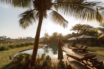 Rear view  young slim woman stand near swimming pool spa under palm tree. Tropic island relax vacation concept